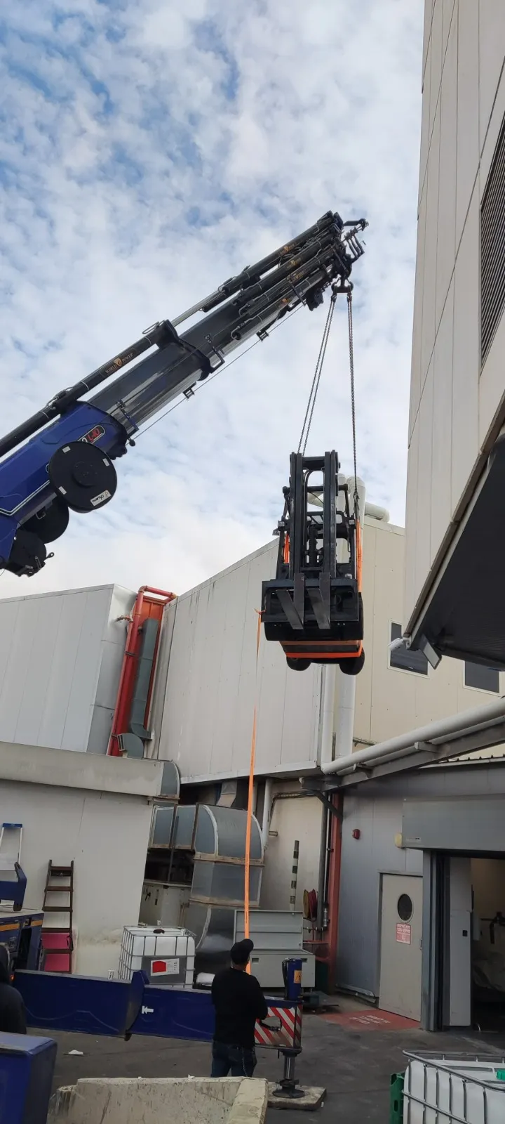 מנוף גדול מרים מלגזה כתומה אל קומה עליונה של מבנה תעשייתי, על רקע שמיים כחולים